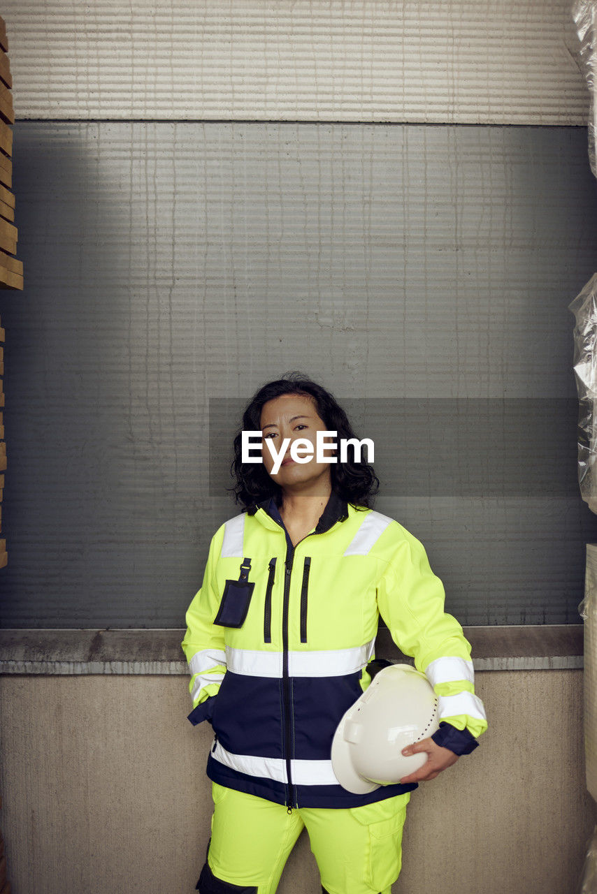 Portrait of confident female worker in reflective clothing standing against wall in factory