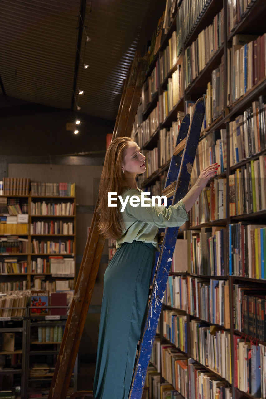 Woman with long hair standing on ladder while searching book in library