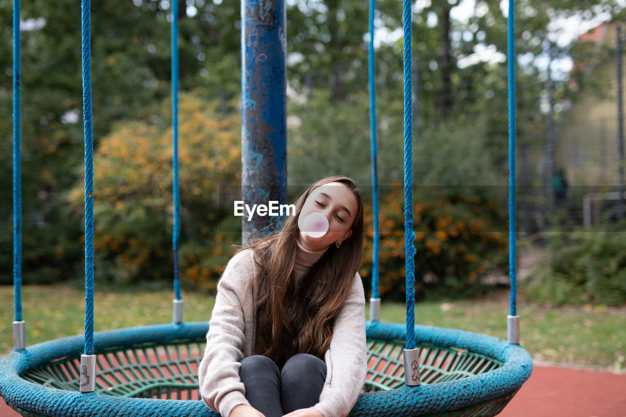 Smiling young woman blowing bubblegum while sitting in swing at playground