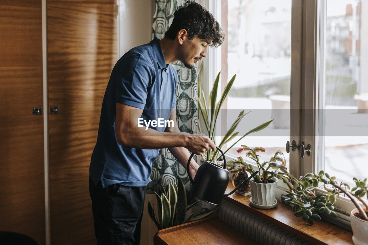 Male care assistant watering plants on window sill at home