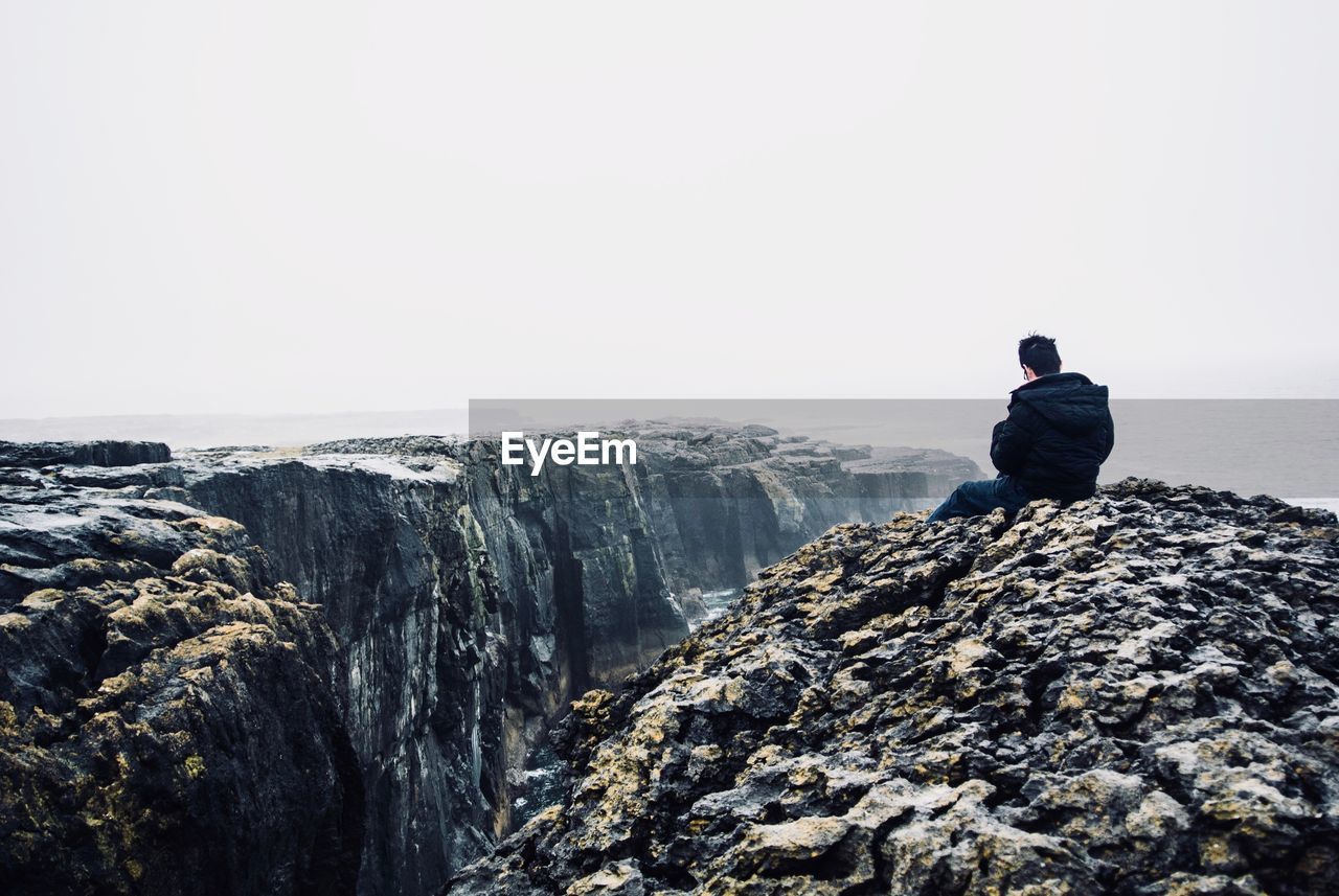 REAR VIEW OF MAN SITTING ON ROCK LOOKING AT VIEW