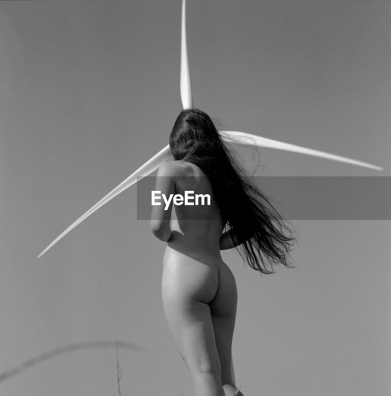 Rear view of naked woman standing by wind turbine against sky