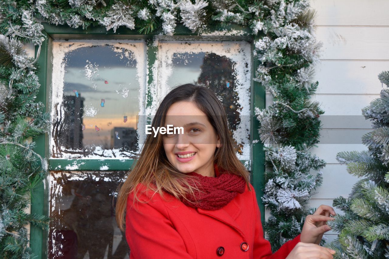 Portrait of smiling young woman standing against decorated window during winter