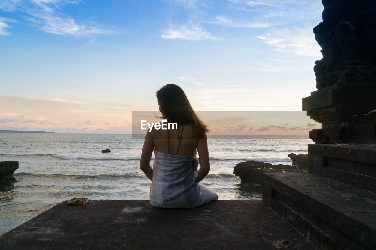 Rear view of woman sitting on wall by sea sky during sunset