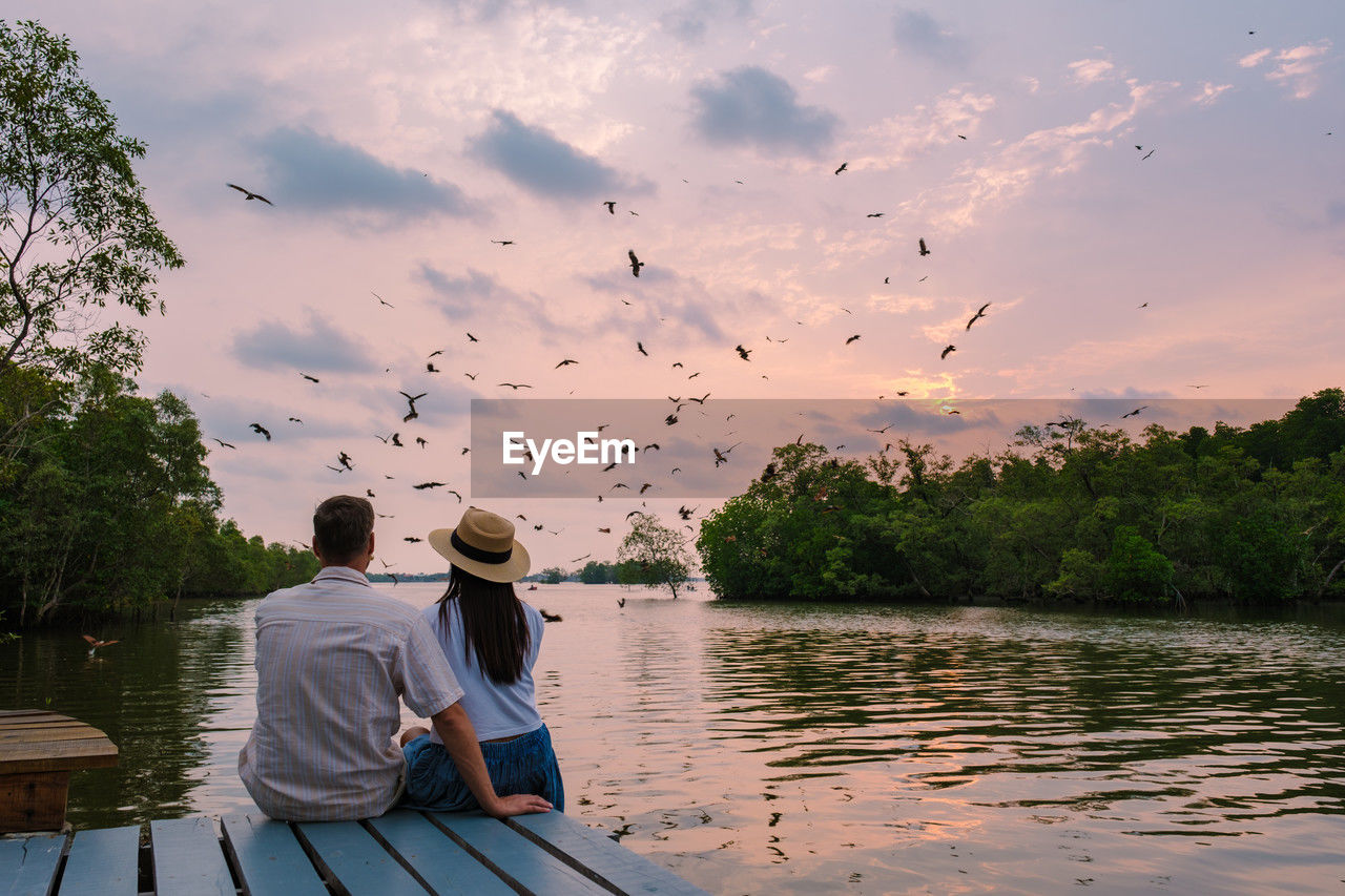 rear view of man sitting on pier against lake against sky during sunset