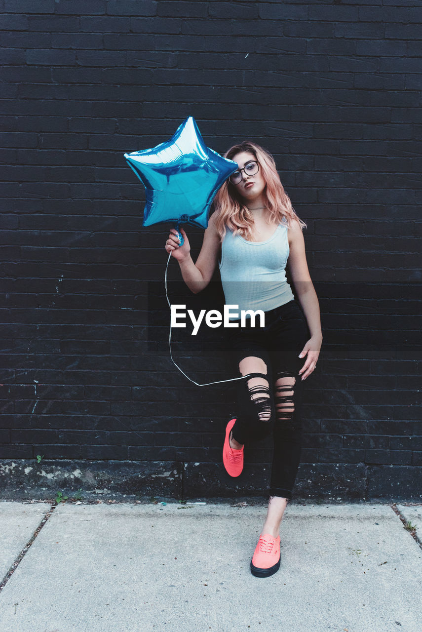 Portrait of woman holding blue star shape balloon while leaning on wall