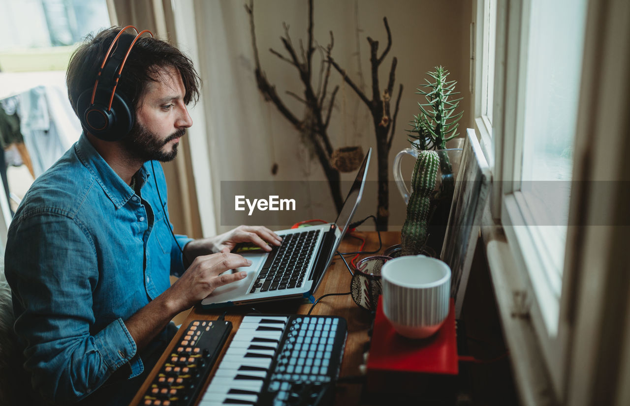 Side view of young man in headphones using synthesizer and laptop at table at home