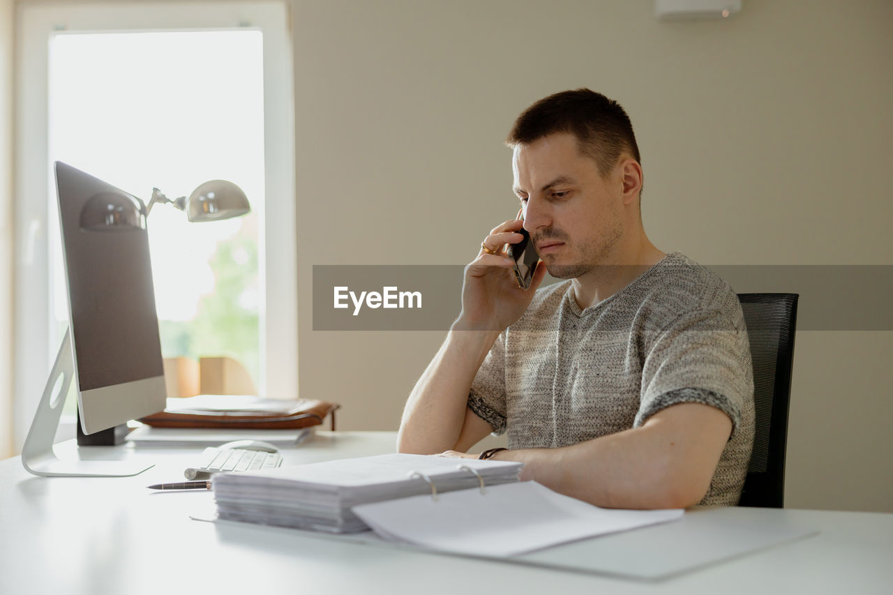 Young caucasian man sitting at his desk in office, working with computer and communicating