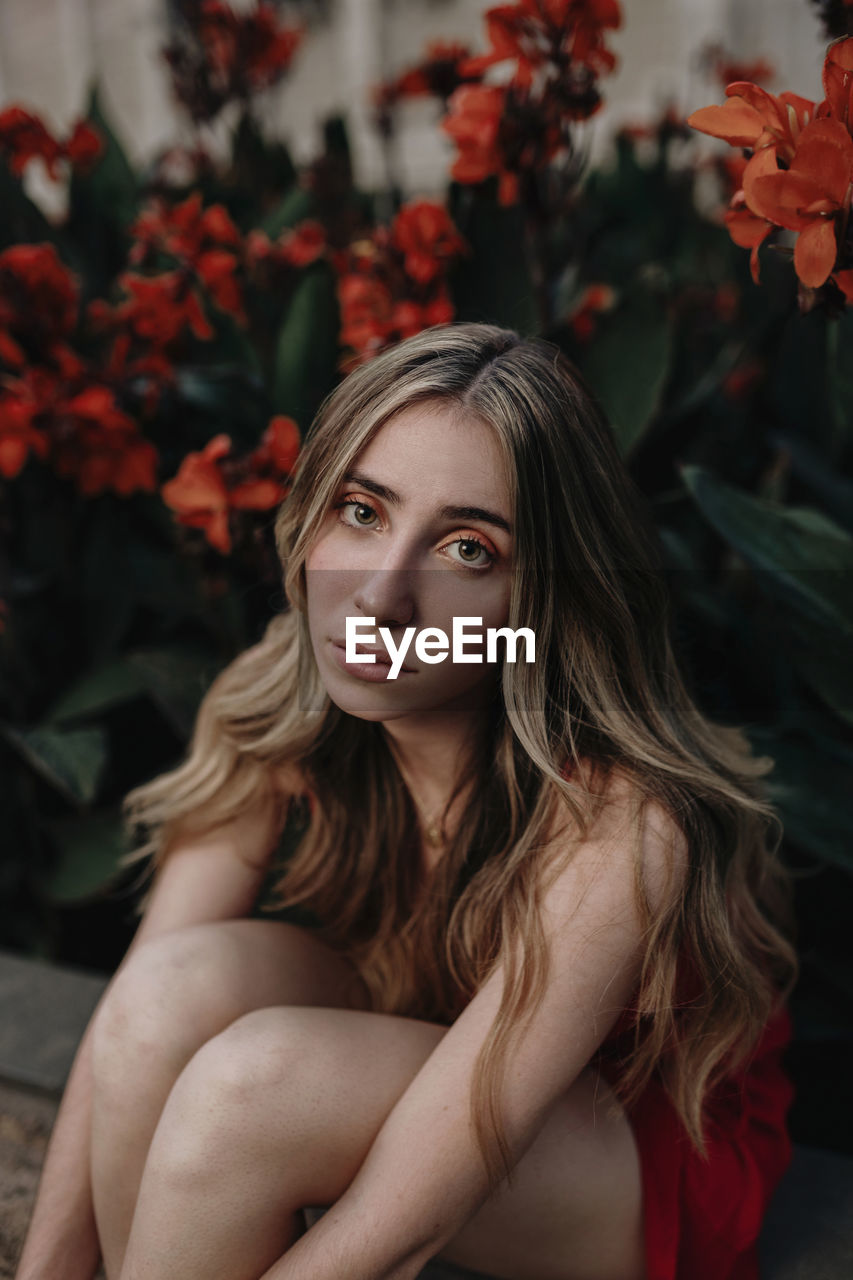 Young mindful female with makeup sitting near blossoming flowers on city street in summer