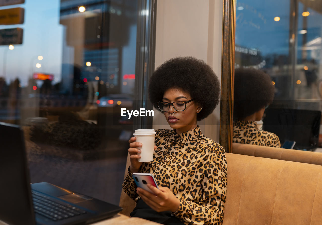 Serious african american female with afro hair scrolling cellphone while working remotely at table with laptop in cafe sitting near glass window enjoying hot beverage on take away cup