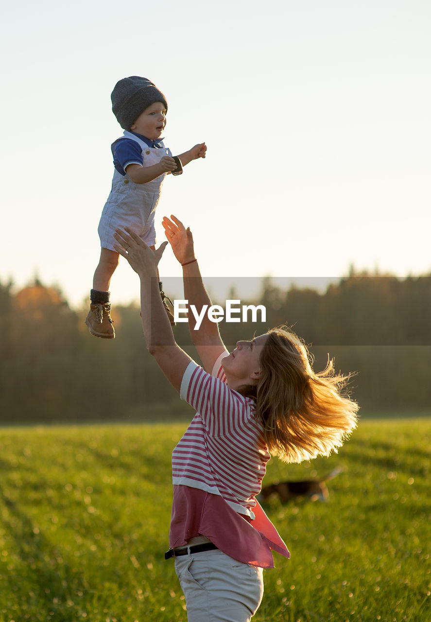 Playful mother catching son on field against sky