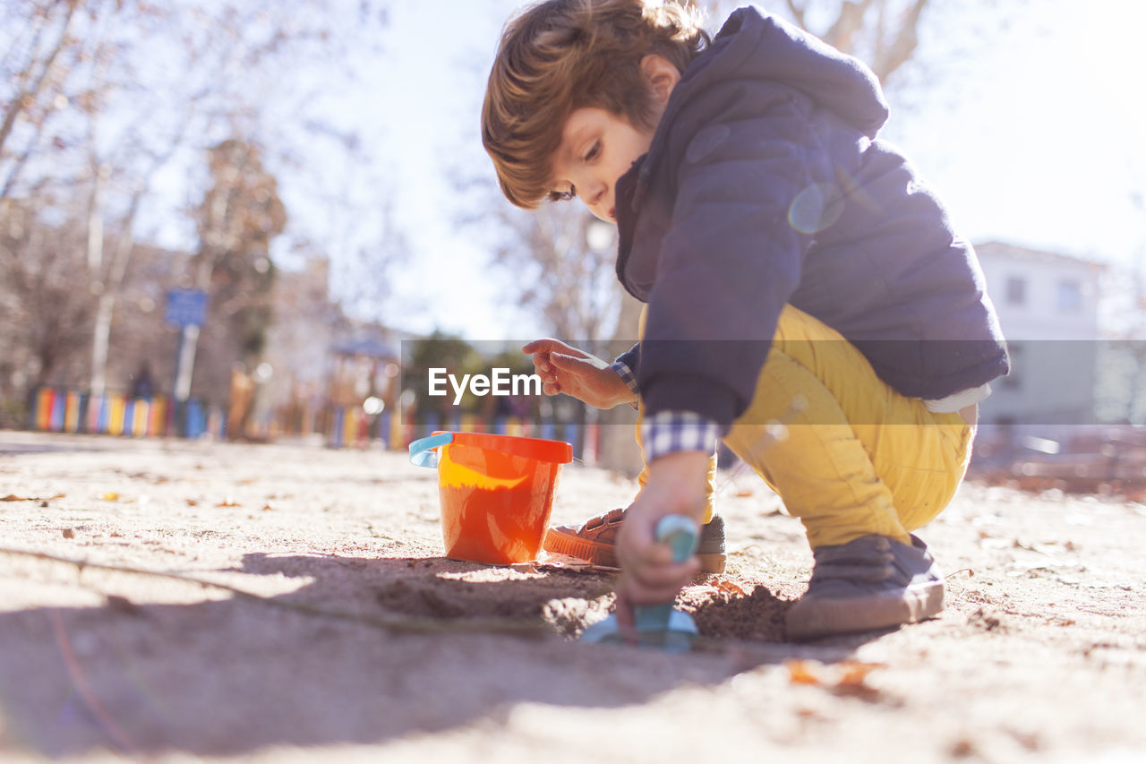 Side view of boy playing with sand pail and shovel while crouching at beach