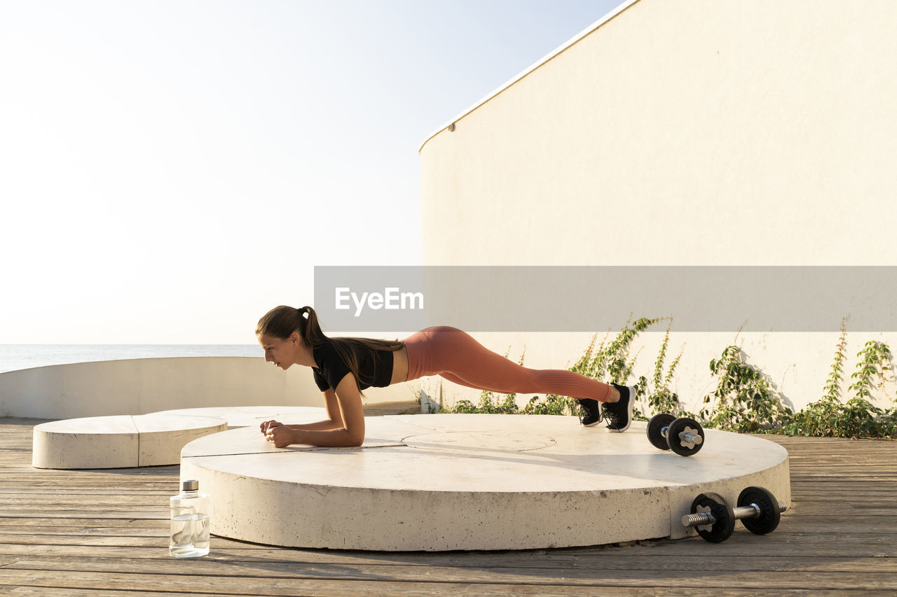 Sportswoman doing plank position exercise on pedestal by sea during sunrise