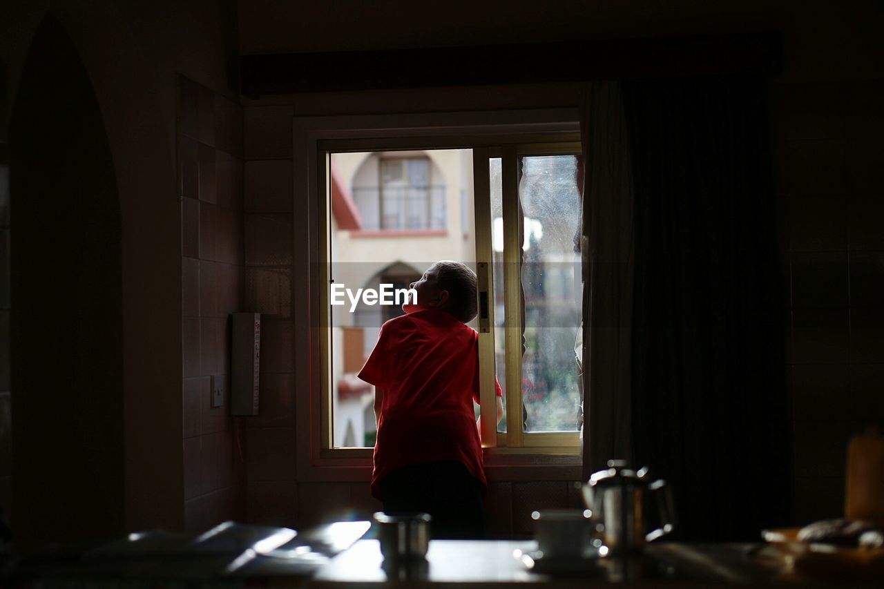 Rear view of boy looking up through window at home