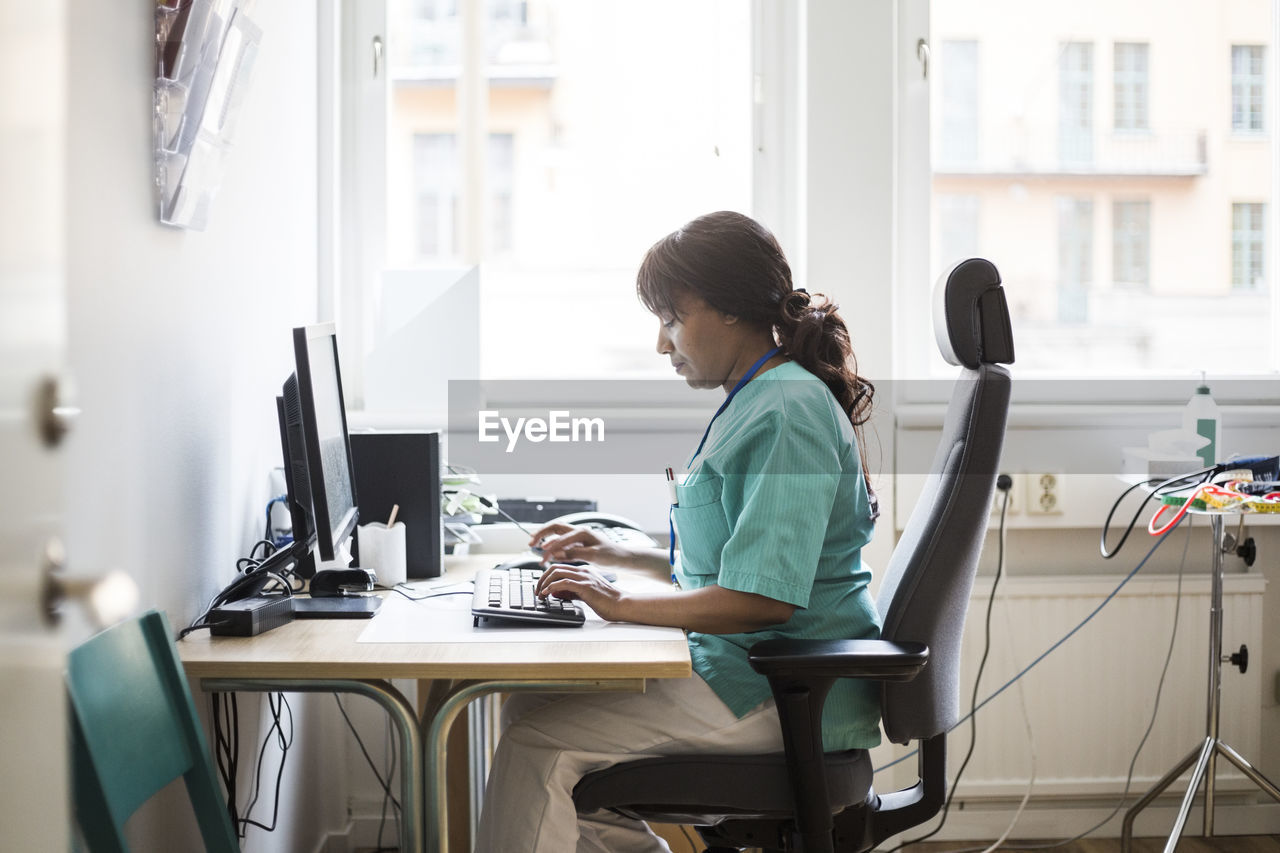 Side view of mature female doctor typing over computer while sitting in clinic