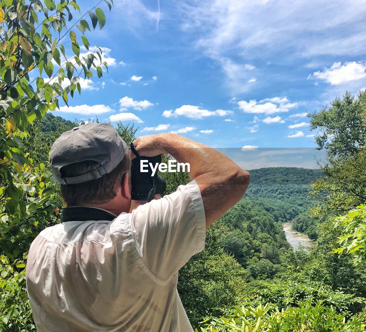 Rear view of senior man photographing with camera while standing on mountain against blue sky during sunny day