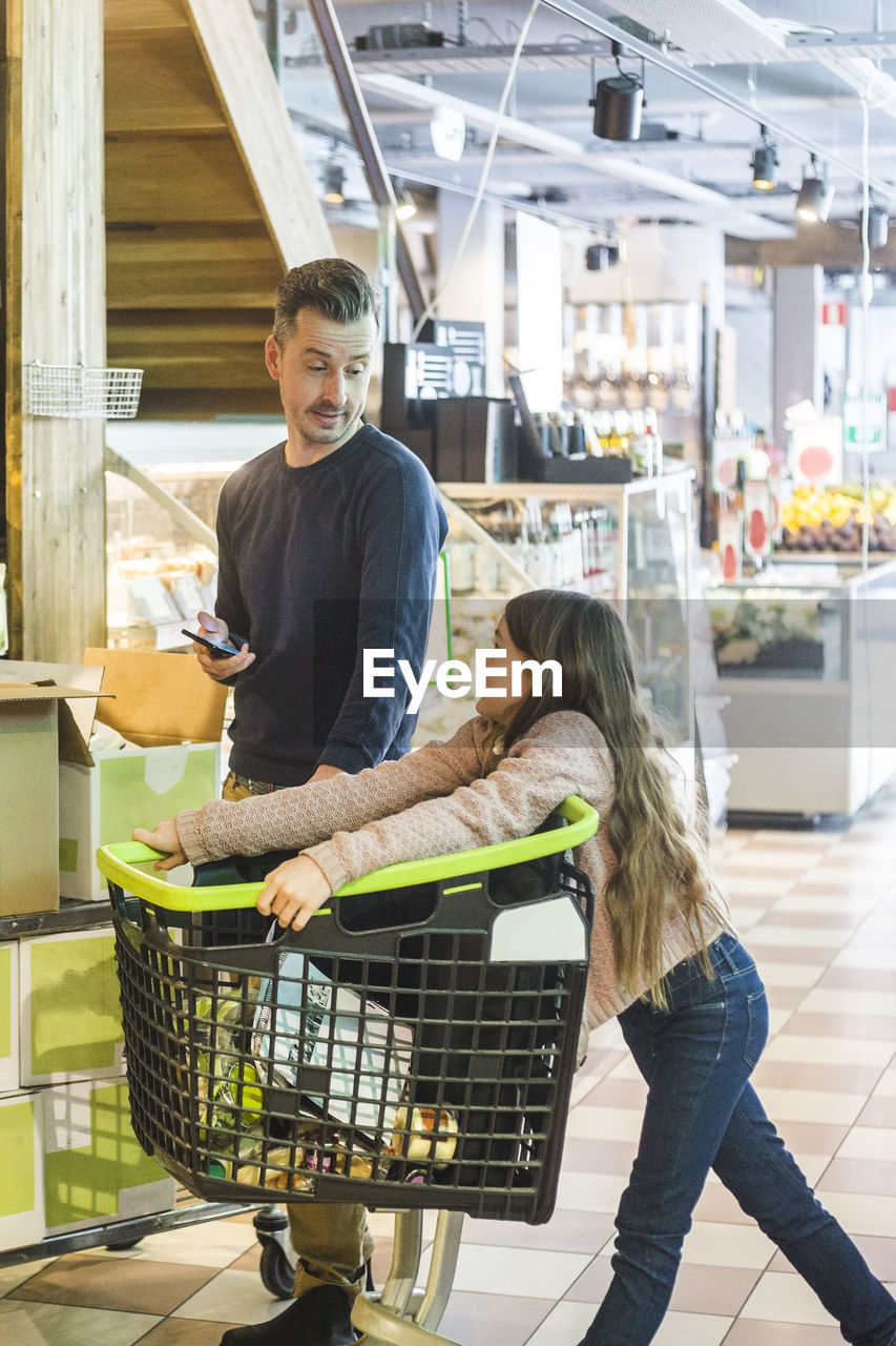 Father talking to cute daughter leaning on shopping cart at supermarket