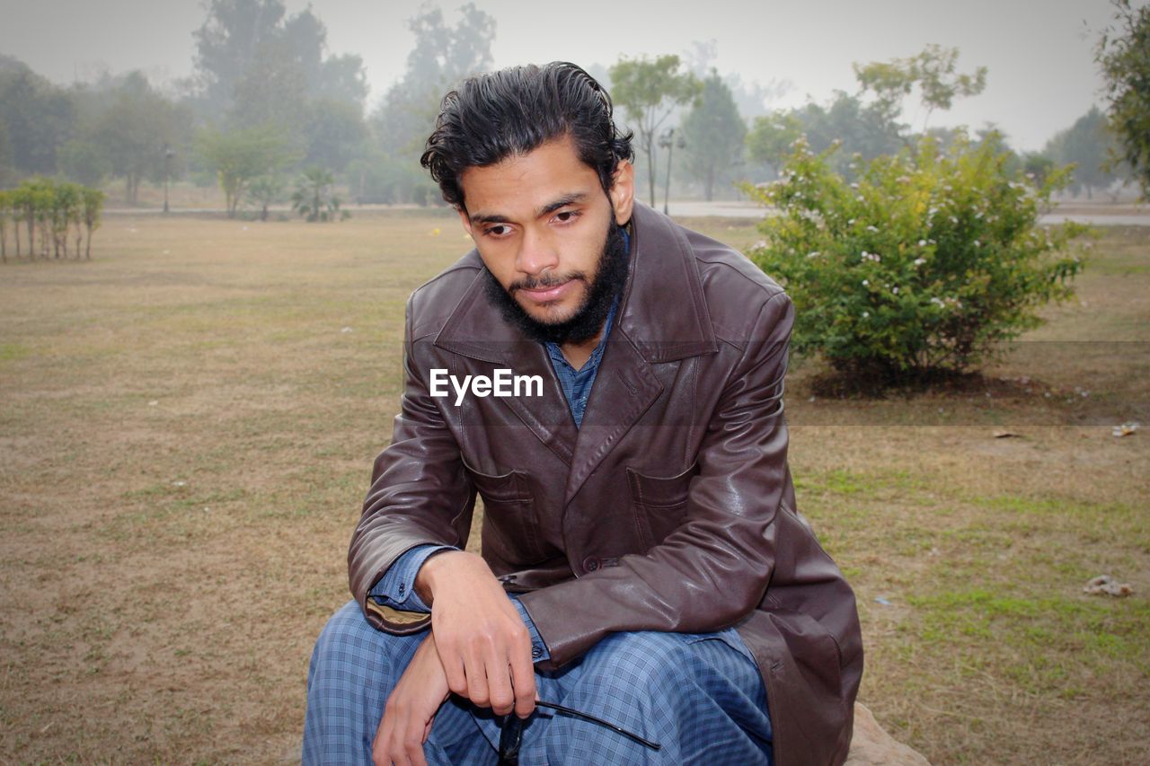 one person, adult, men, sitting, portrait, young adult, looking at camera, person, front view, nature, beard, casual clothing, three quarter length, lifestyles, plant, leisure activity, facial hair, serious, clothing, relaxation, emotion, photo shoot, outdoors, human face, looking, jeans, day, copy space, tree, black hair, contemplation