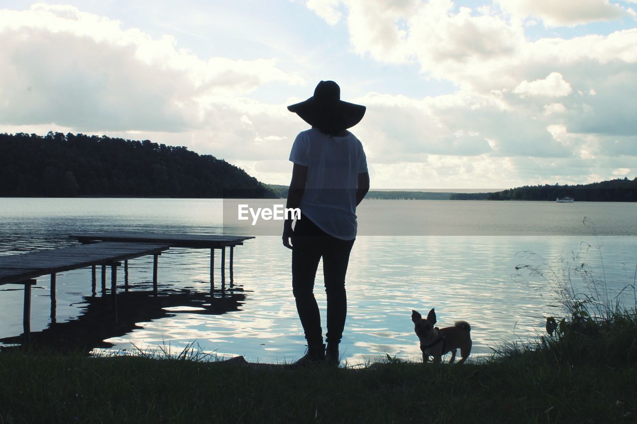 Rear view of woman wearing hat standing with dog at riverbank against sky