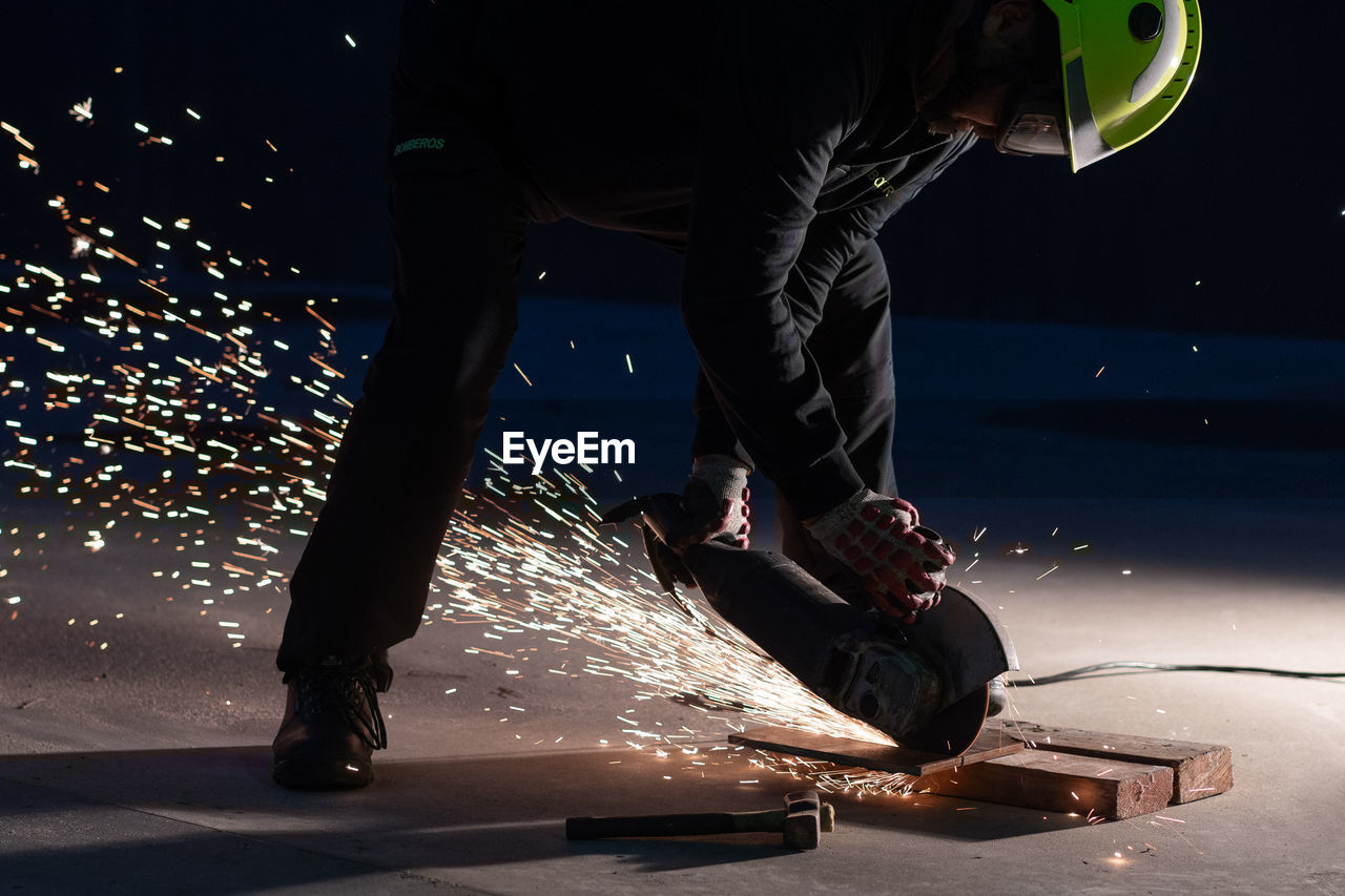 Unrecognizable male welder in protective helmet and goggles cutting iron details with chop saw during work at industrial factory at night