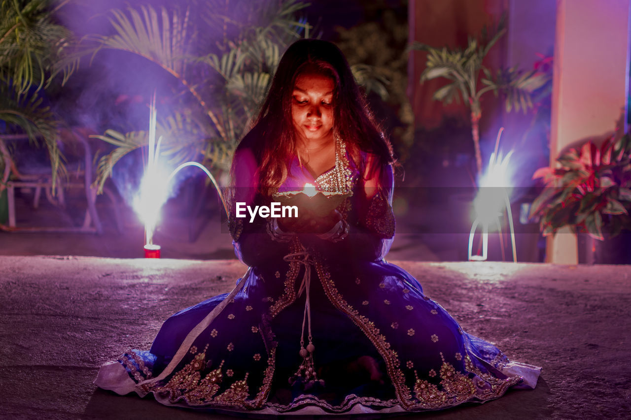 Young woman holding diya while sitting outdoors at night