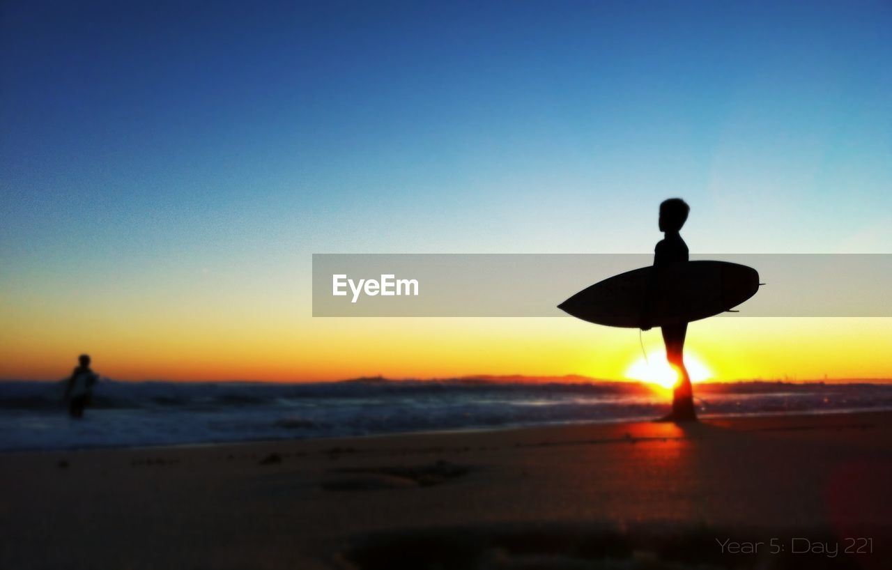 Silhouette boy with surfboard standing at shore on beach during sunset