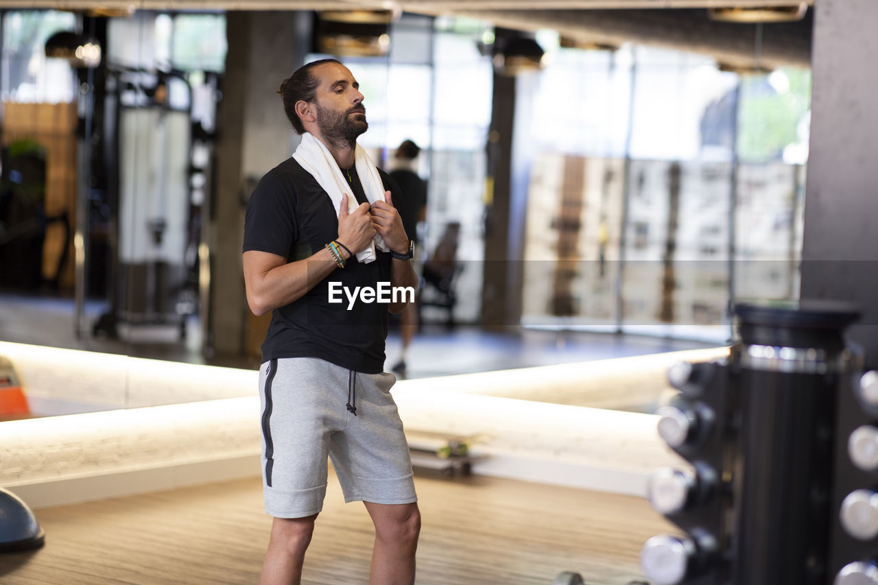 Exhausted bearded male athlete with towel closing eyes and resting after fitness workout in modern gym