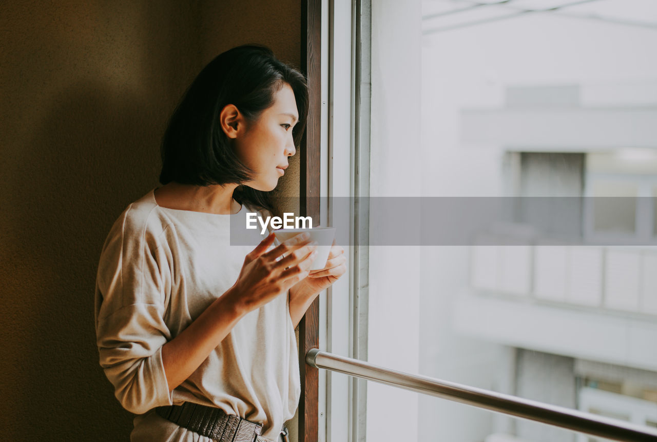 YOUNG WOMAN DRINKING COFFEE FROM WINDOW AT HOME