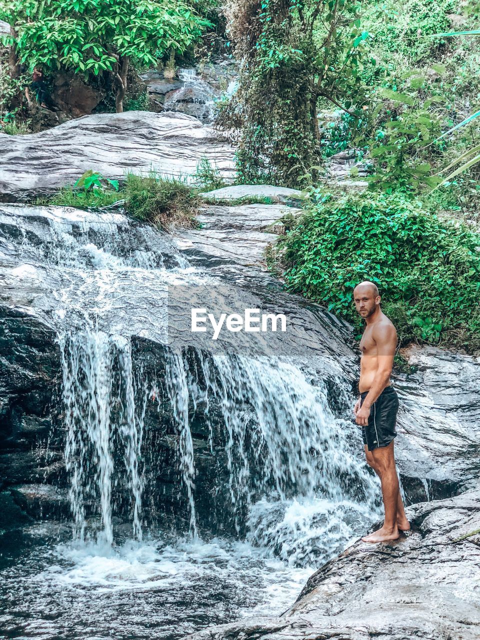 Portrait of shirtless man standing by waterfall in forest