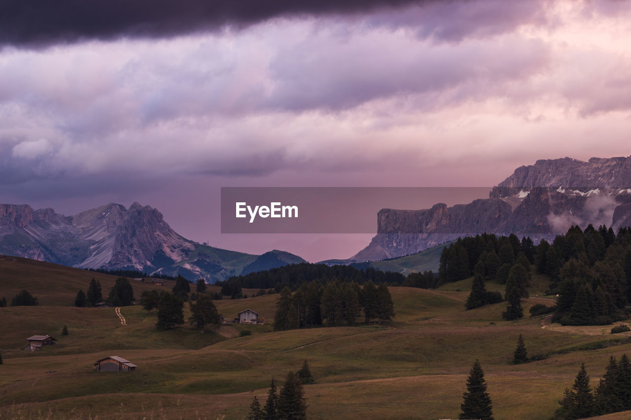 Meadow in the mountains with trees and cabins against dark cloudy sky during sunset