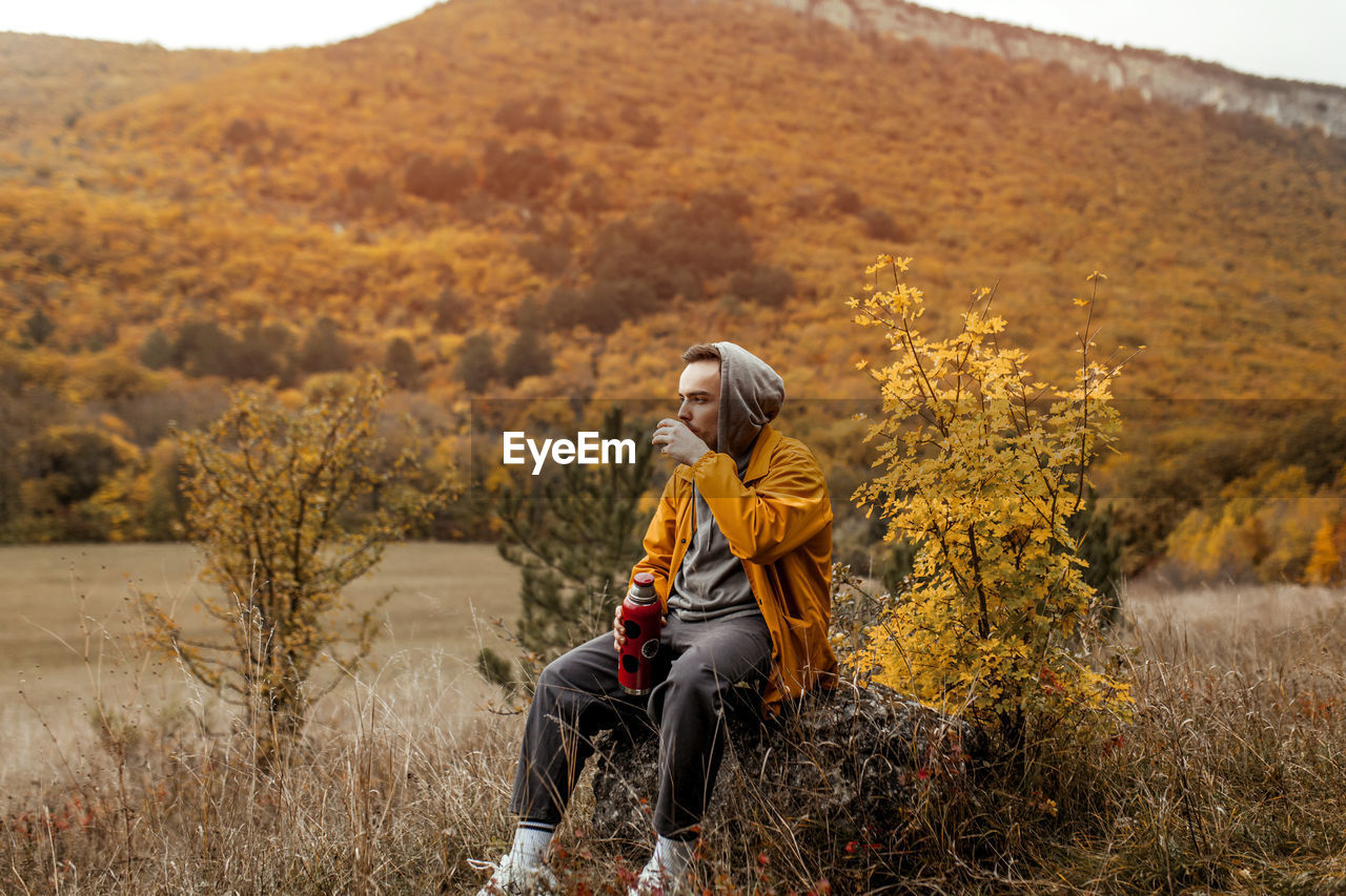 A young man in a yellow jacket pours and drinks tea from a thermos