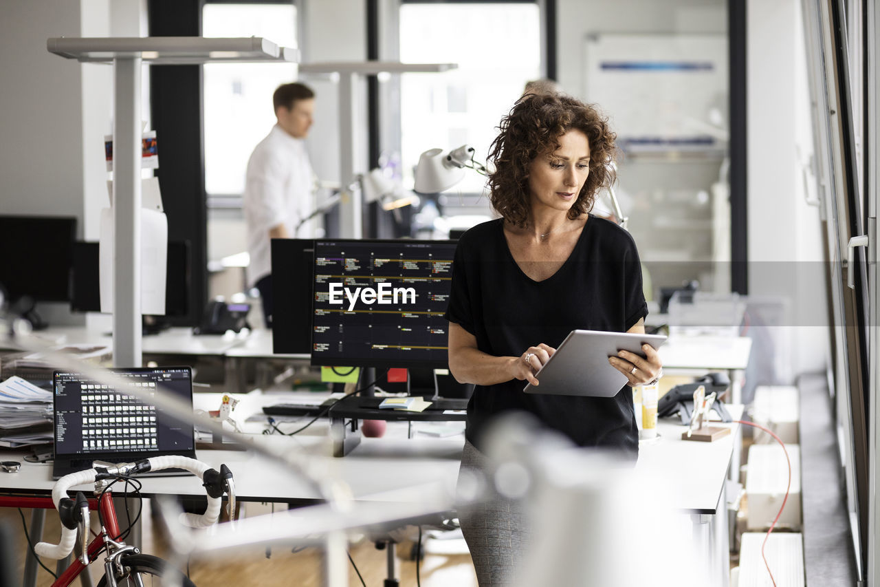 Businesswoman using digital tablet with colleague standing in background at open plan office