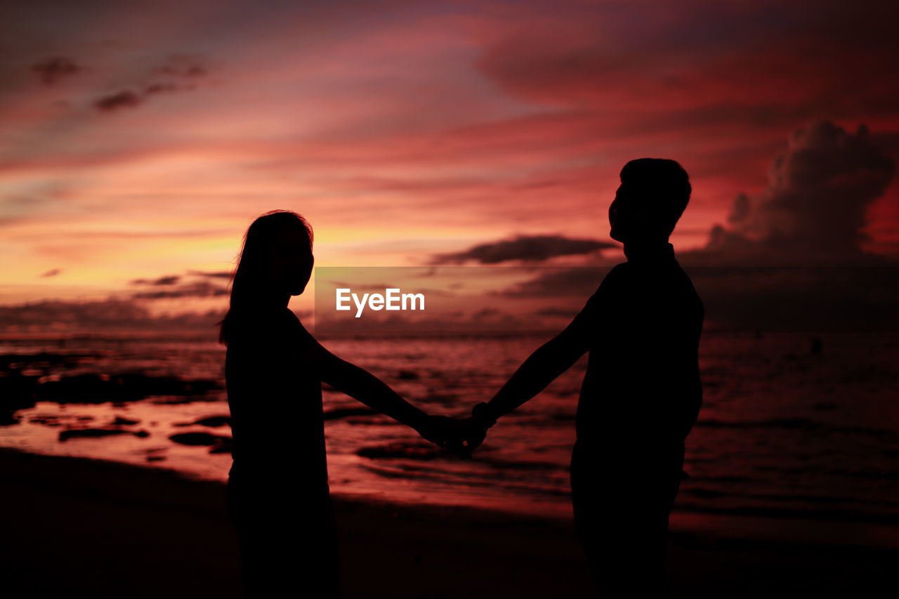 Silhouette couple holding hands while standing on beach during sunset