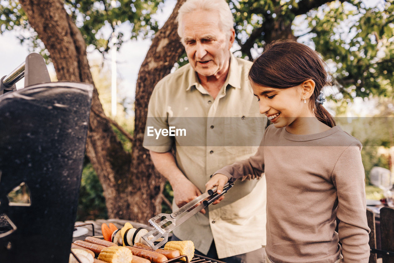 Smiling girl barbecuing with grandfather in back yard at dinner party