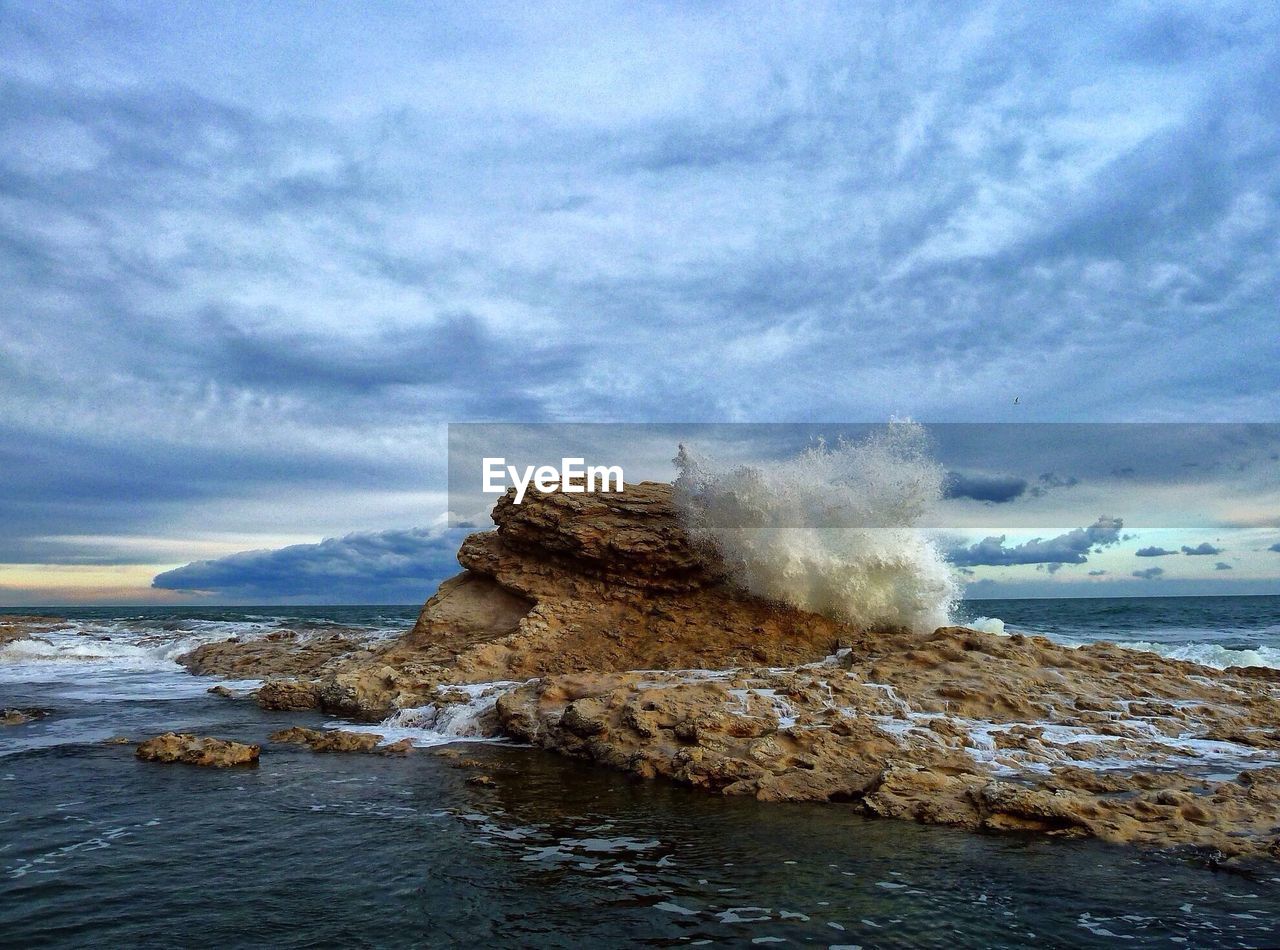 Waves splashing on rocks against cloudy sky