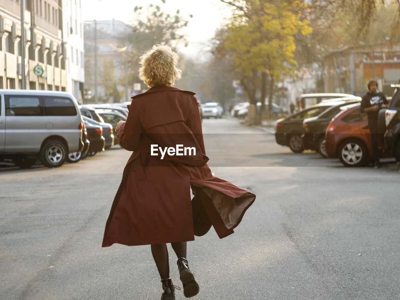 Young woman with curly hair walking on city street at sunset wearing red coat