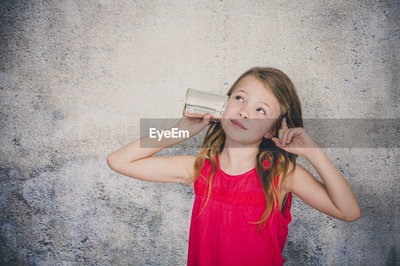 Cute girl playing with tin can phone while standing against wall