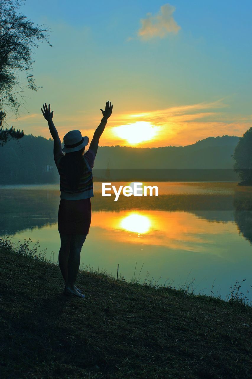 Rear view of woman standing by lake against sky during sunset