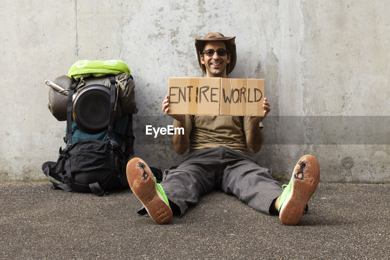 Portrait of man holding cardboard with entire world text while sitting against wall 