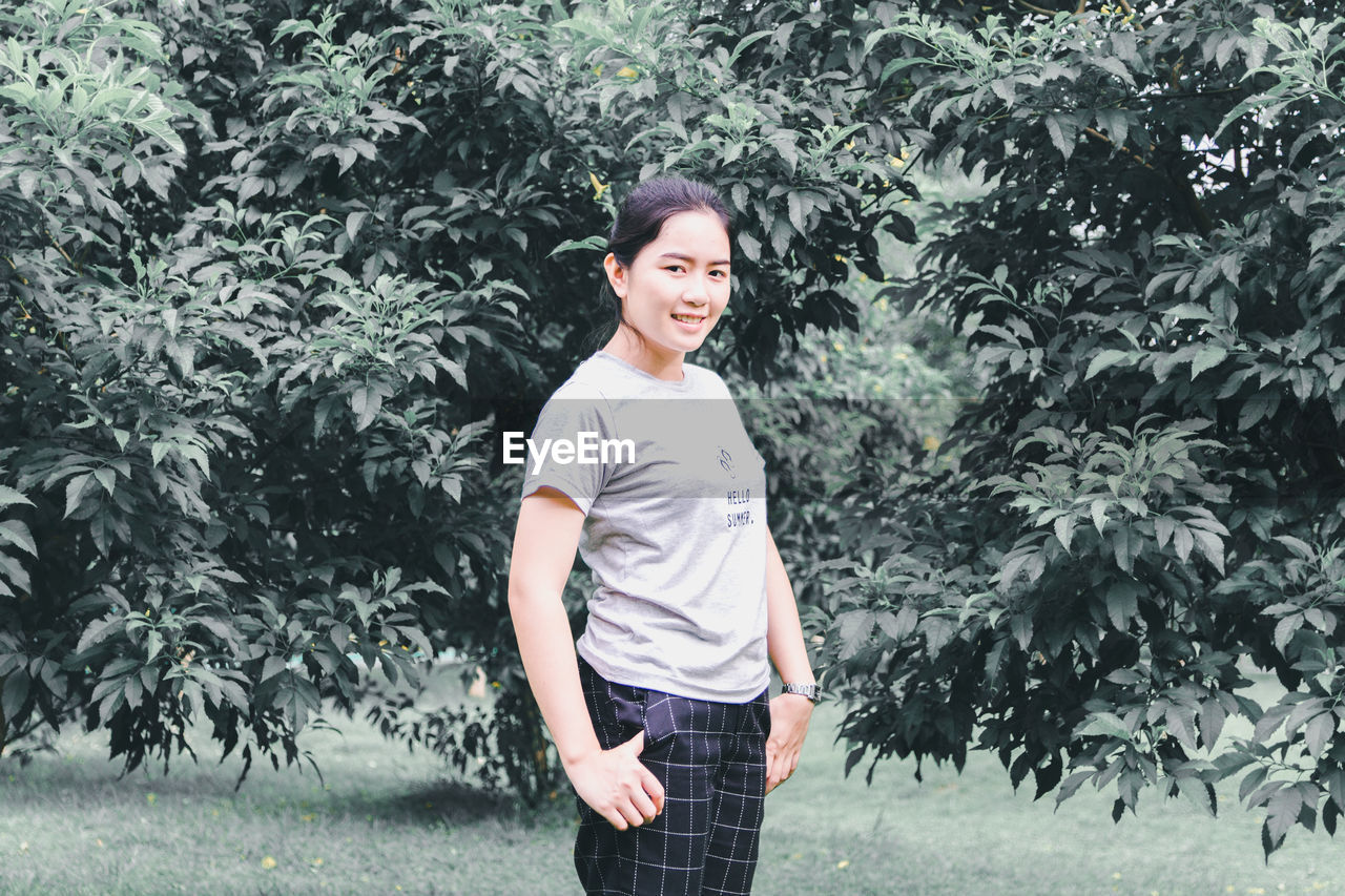 PORTRAIT OF A SMILING YOUNG WOMAN STANDING AGAINST PLANTS