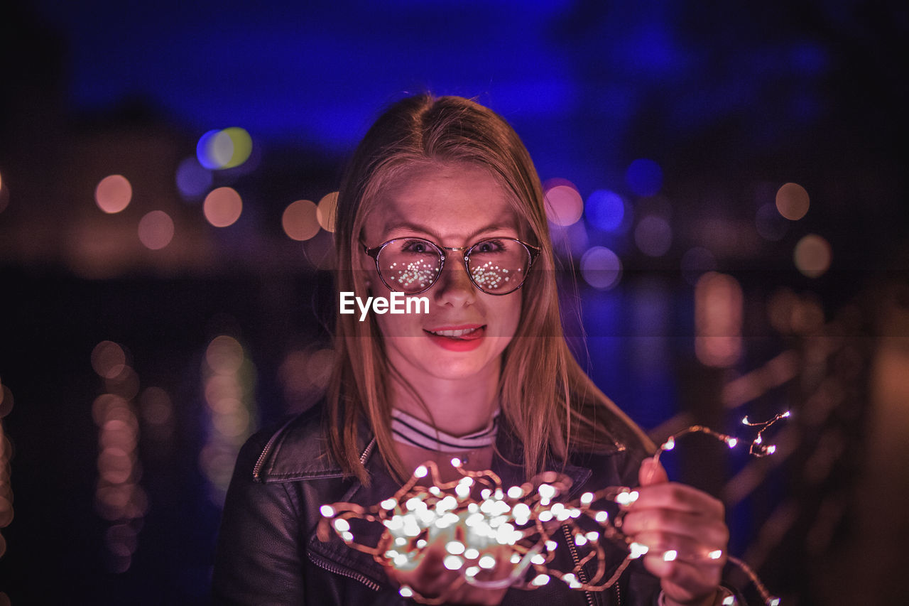Portrait of young woman wearing eyeglasses holding illuminated string lights in city at night