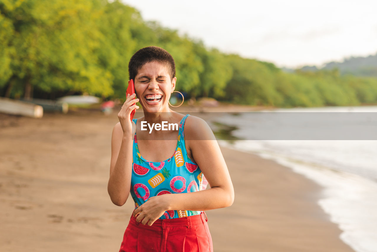 Cheerful young latin american woman with short hairstyle talking on mobile phone while standing with eyes closed near waving sea in summer day