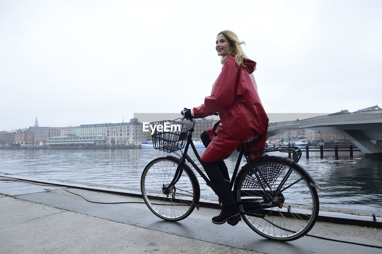 Denmark, copenhagen, happy woman riding bicycle at the waterfront in rainy weather