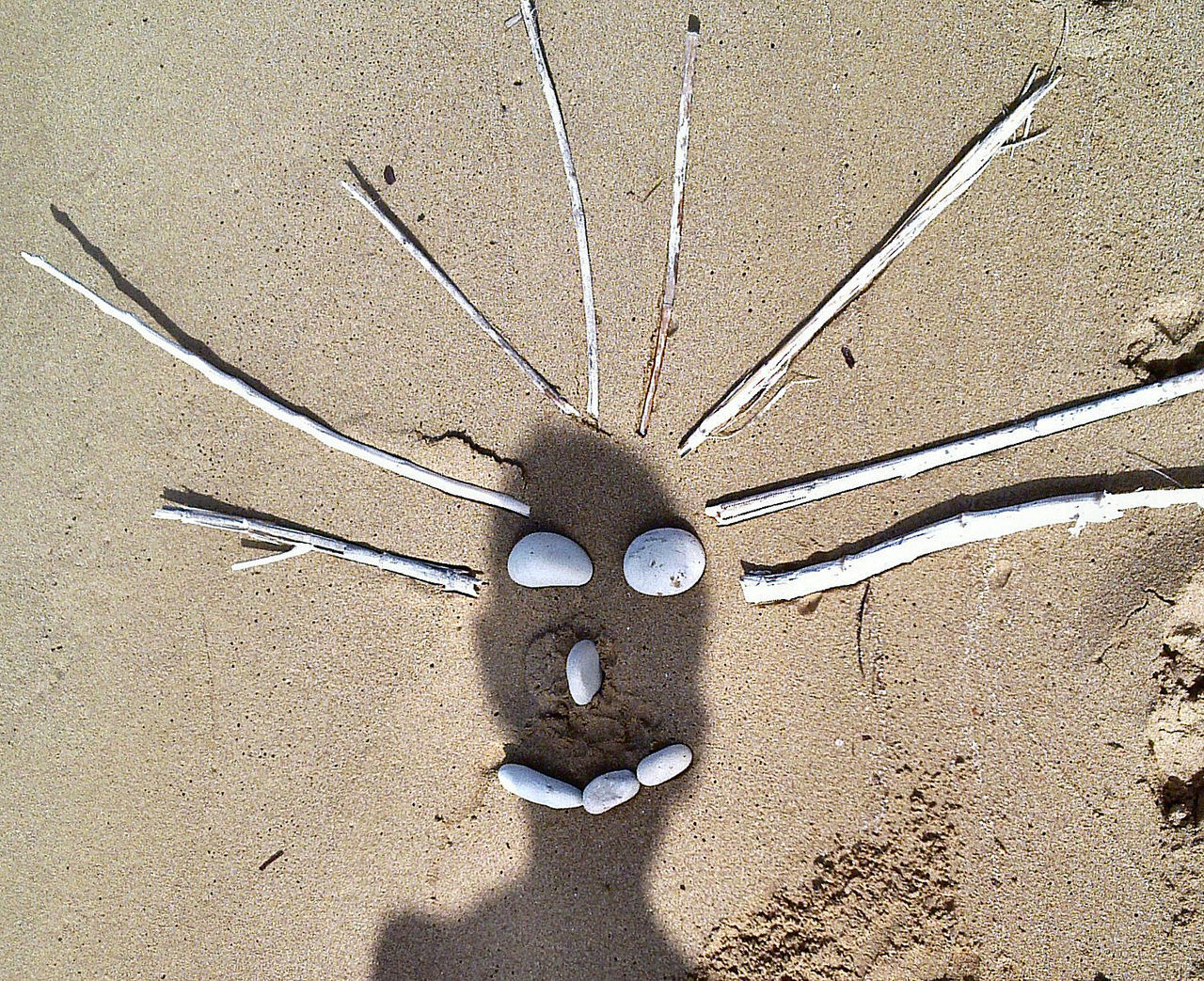 High angle view of person shadow on anthropomorphic face made from pebbles and sticks at beach