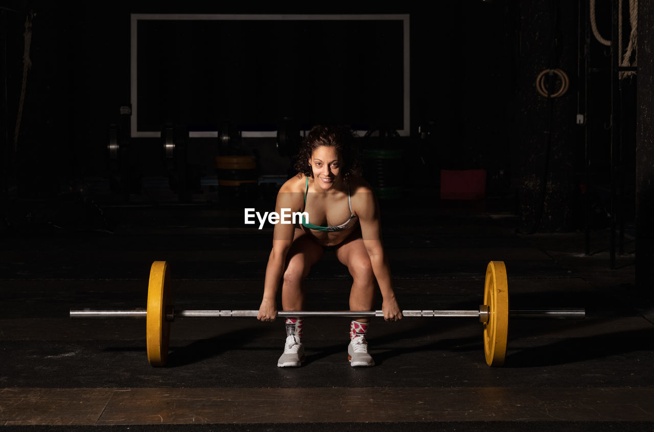 Happy muscular woman win sportswear smiling and lifting heavy barbell over head during intense workout in dark gym