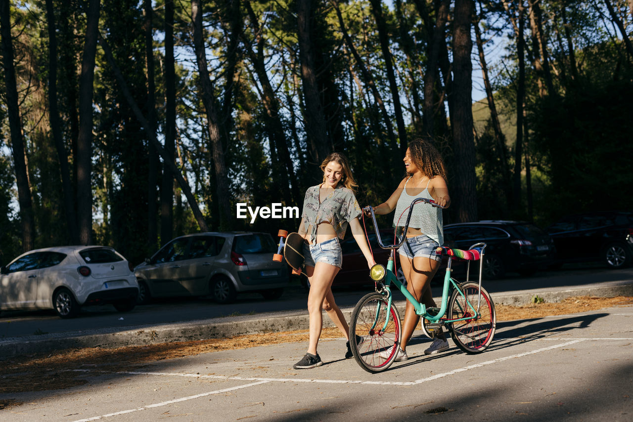 Female friends walking on street holding skate board and bicycle in vacations