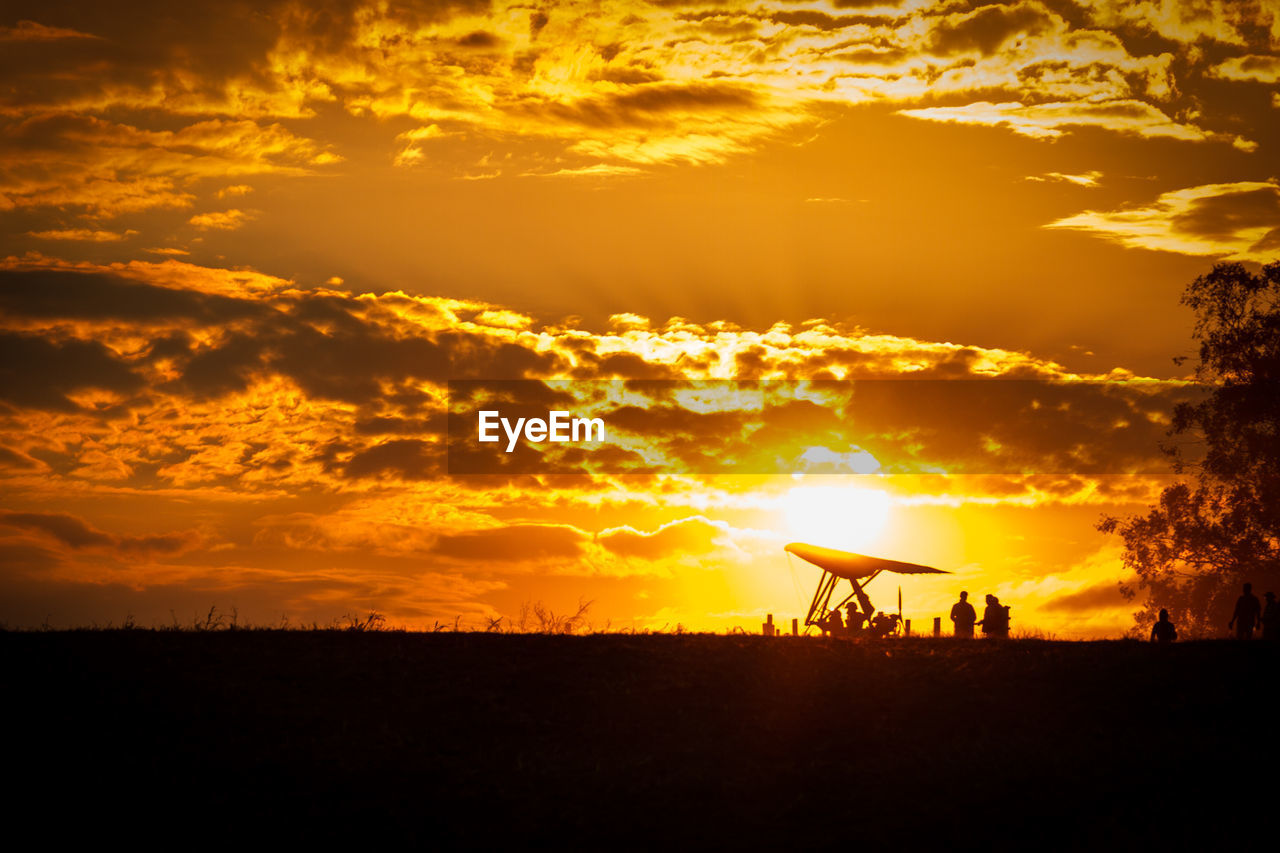 People preparing for hang-gliding on silhouette field against dramatic sky during sunset