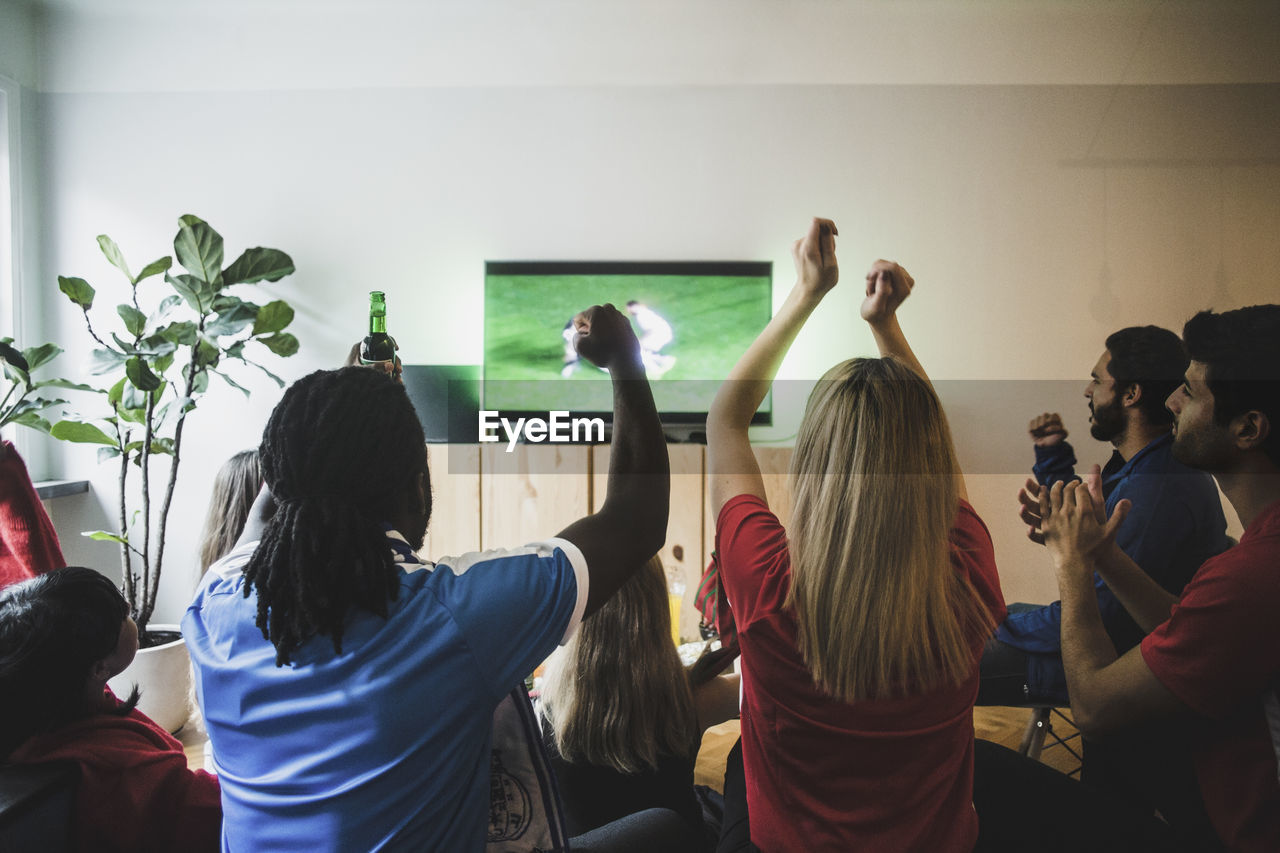 Cheerful friends with arms raised watching soccer match in living room