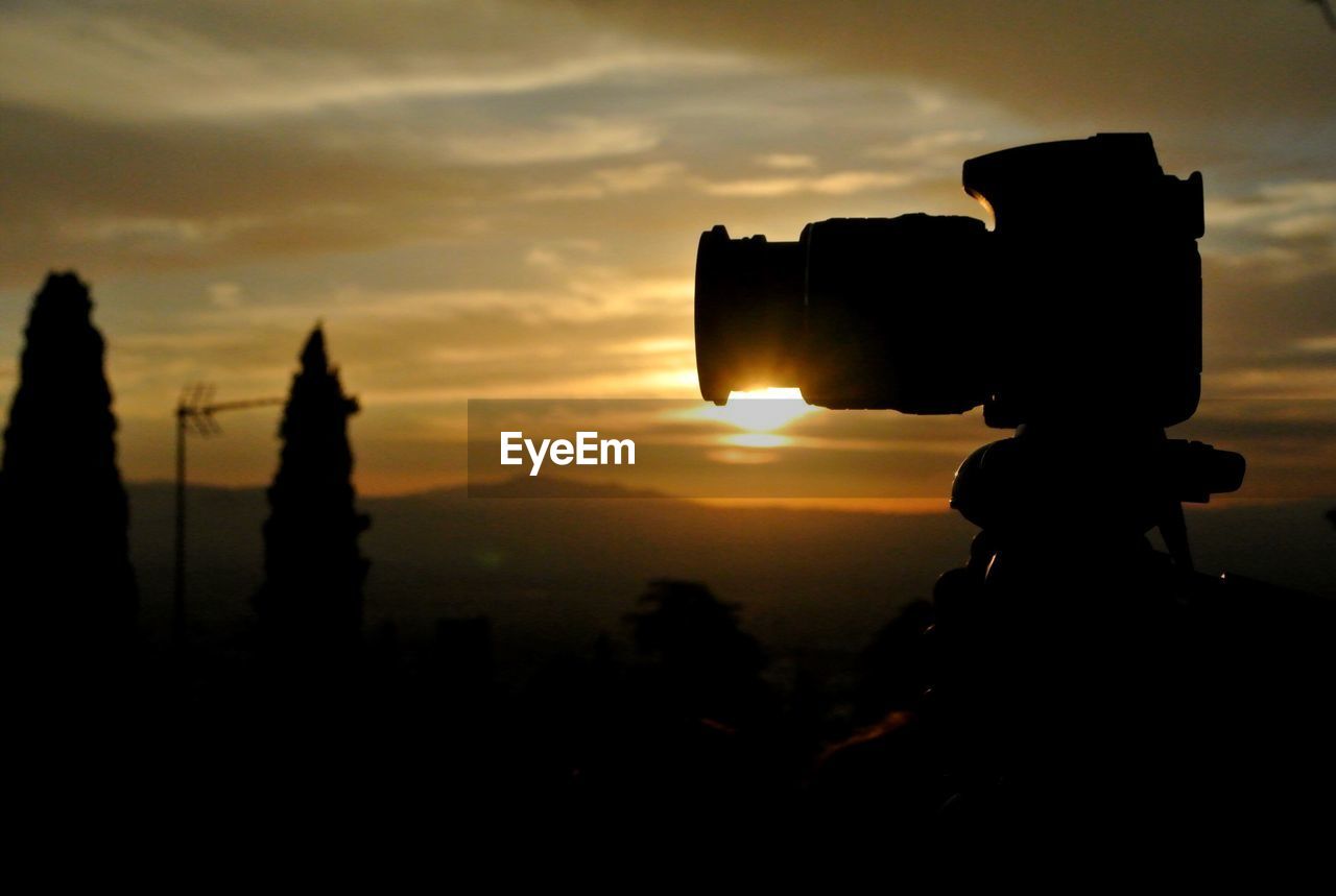 Close-up of back lit camera on tripod against sky during sunset