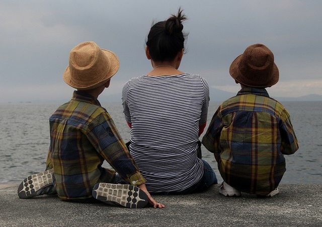 Rear view of mother with children sitting on retaining wall by sea against cloudy sky